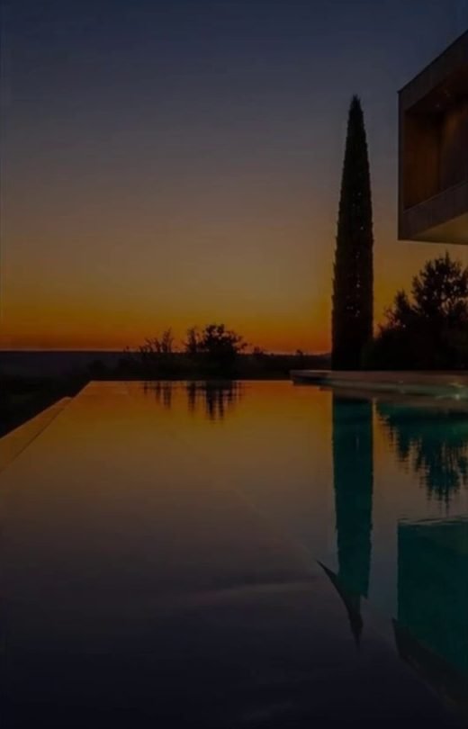 Piscina a sfioro al tramonto con un cipresso e una villa moderna